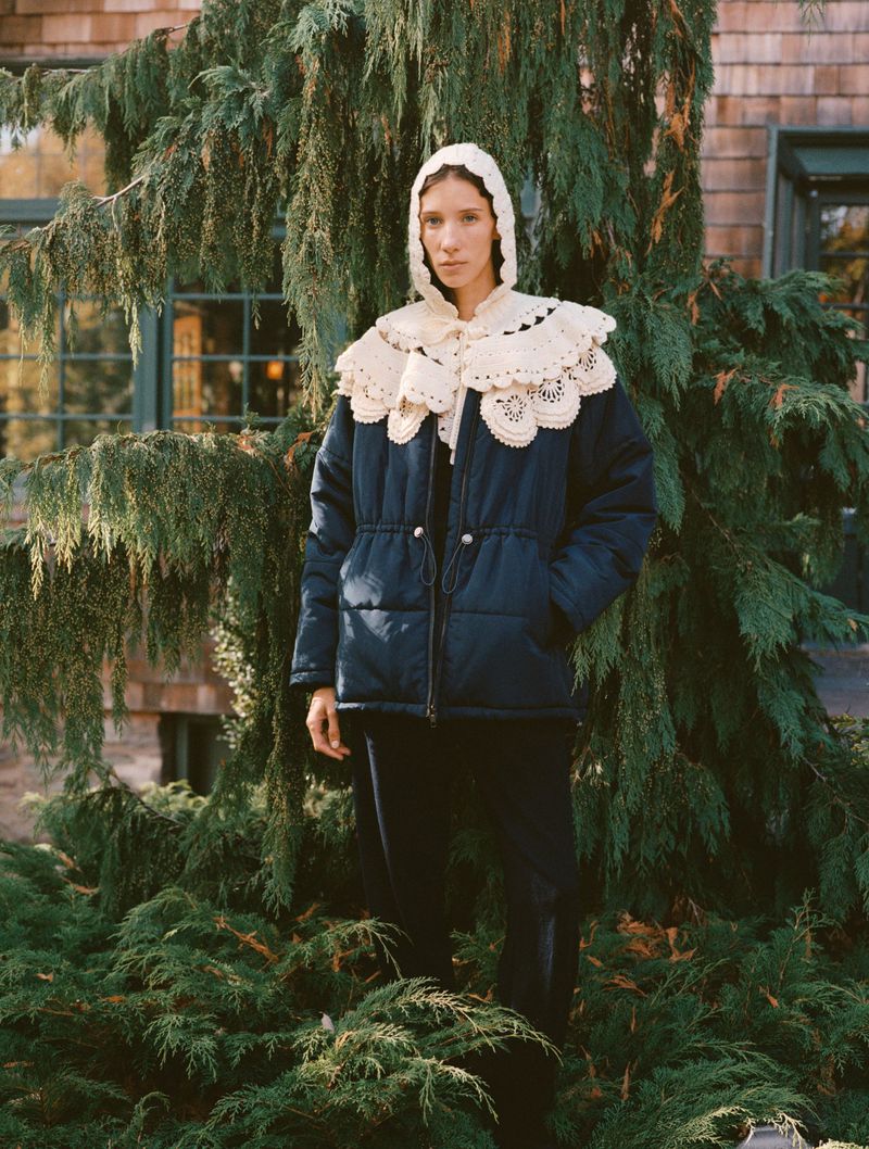 Woman in hooded crochet collar and navy puffer jacket posing outdoors against green bushes and the wall of a light brown house.