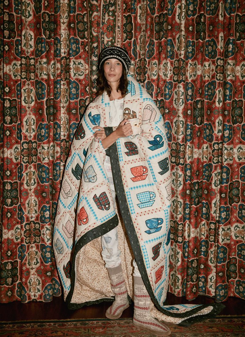 Adult female wrapped in a patchwork quilt with teacup patterns, wearing patterned socks and a hat, standing indoors with patterned wallpaper.