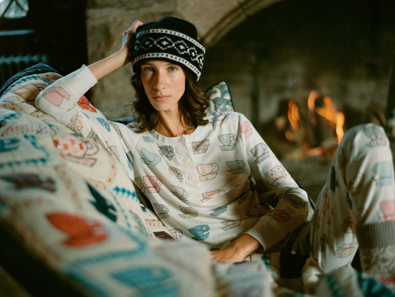 Woman lounges in pajamas with a cup pattern, wearing a patterned beanie, with a lit fireplace in the background creating a cozy indoor scene.
