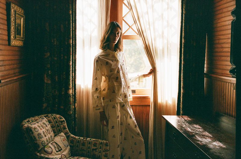 Adult female standing in front of window in white patterned pajama set next to upholstered arm chair inside wood paneled room.