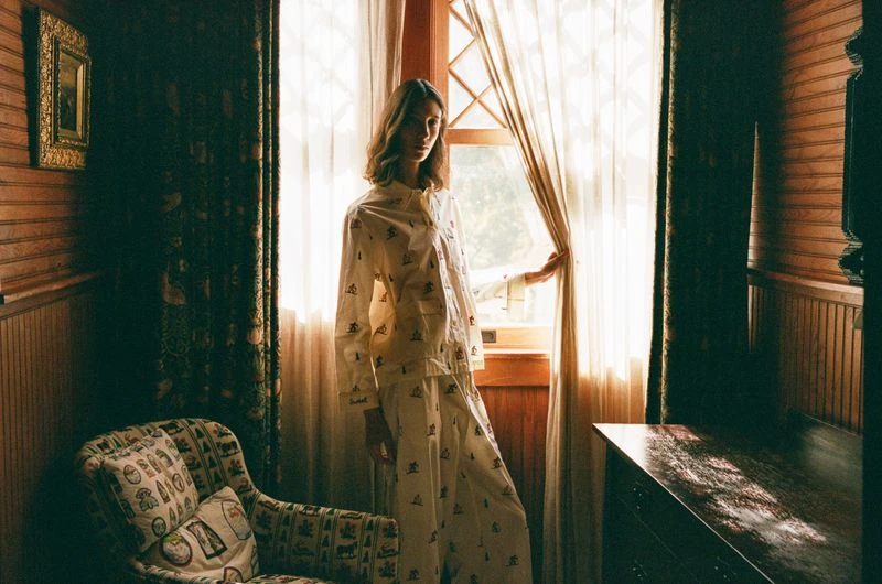 Adult female standing in front of window in white patterned pajama set next to upholstered arm chair inside wood paneled room.