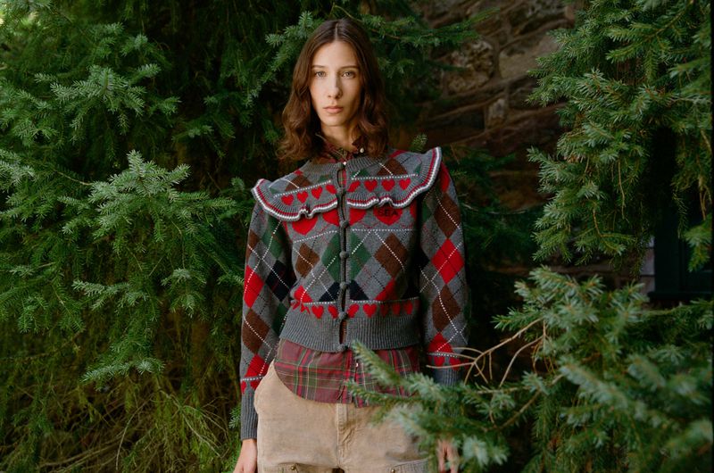 A brunette woman poses outdoors wearing a grey argyle cardigan with red heart details, with a plaid shirt and tan shorts.