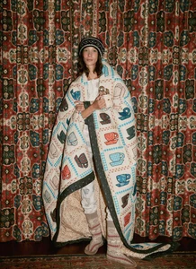 Model Sasha Knysh wrapped in an illustrated quilted blanket wearing a knit headband and patterned socks against tapestry curtains.
