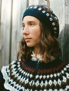 Model Sasha Knysh wearing a navy patterned knit beret and fair isle zip-up sweater against a rustic wooden wall.