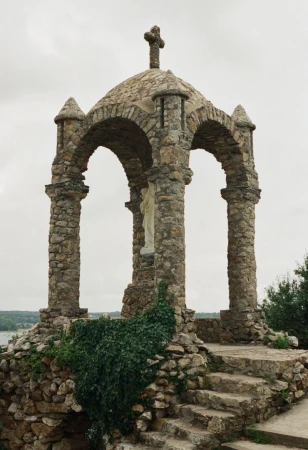 Stone religious shrine with cross on top, arches and pillars, steps in outdoor setting with foliage against cloudy sky background.
