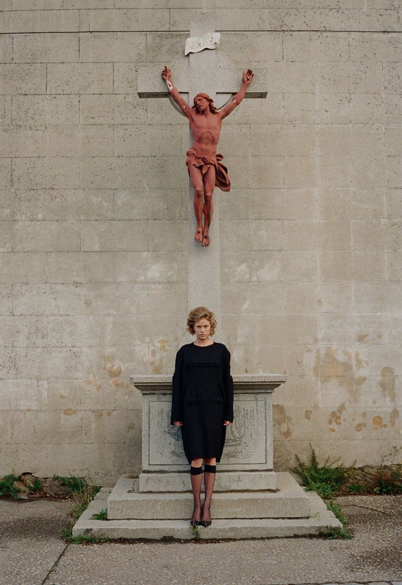A woman in a black dress stands on a stone platform beneath a crucifix statue on a weathered wall in an outdoor setting.