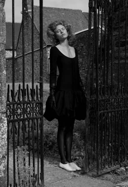 A woman in a black dress poses by an iron gate with a stone building in the background; the image is shot in black and white.