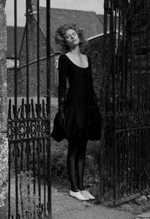 A woman in a black dress poses by an iron gate with a stone building in the background; the image is shot in black and white.