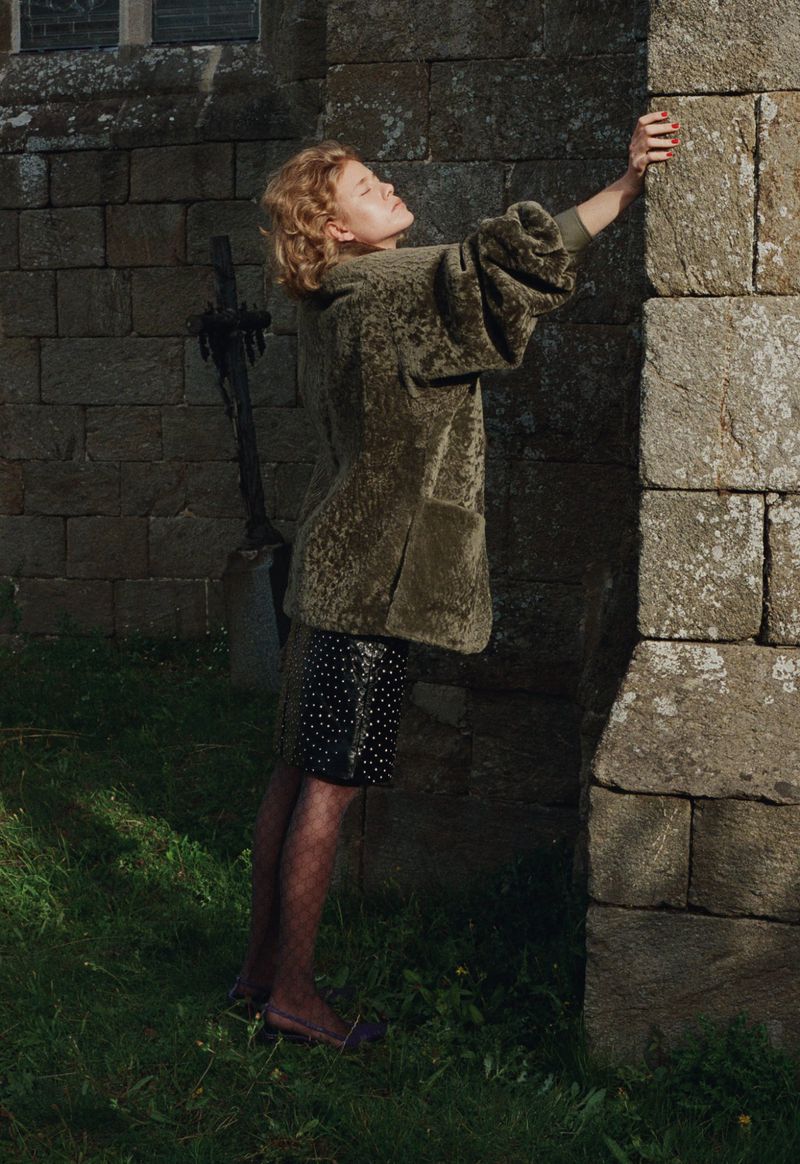 Woman poses by a stone church wall, wearing an olive velvet jacket and polka dot skirt on green grass in soft outdoor light.