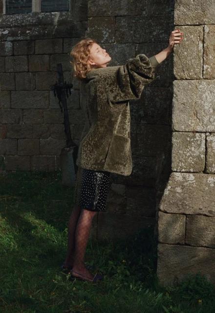 Woman poses by a stone church wall, wearing an olive velvet jacket and polka dot skirt on green grass in soft outdoor light.