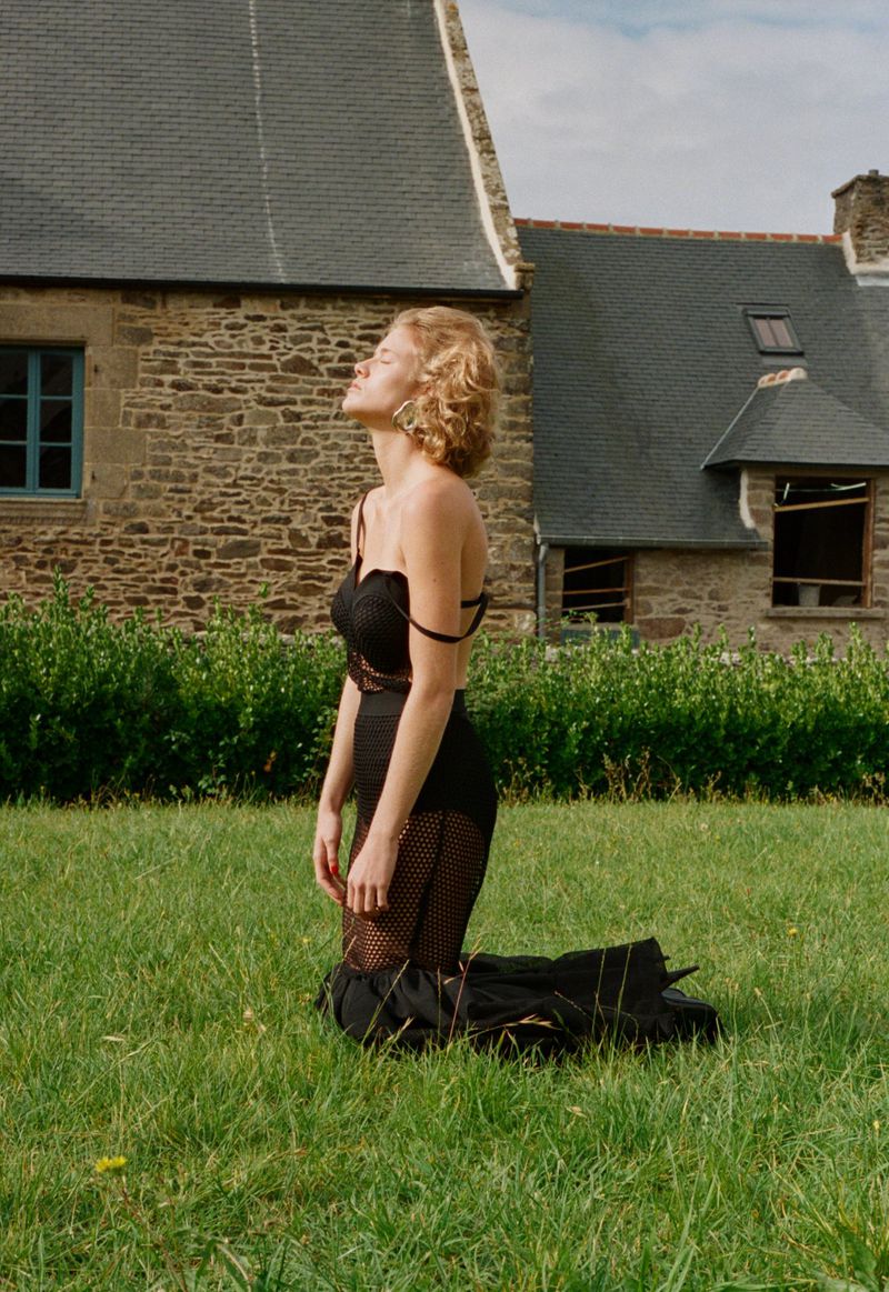 Young woman in black dress kneels in grassy field in front of stone house with eyes closed, soaking in the sun, looking peaceful.