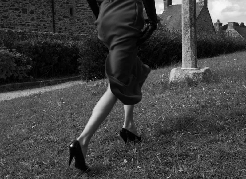 A black and white shot of a woman walking uphill on a grassy field, wearing a dress and high heels, with buildings in the background.