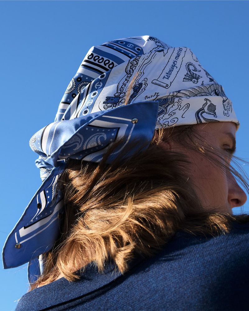 Woman wearing a blue and white patterned headscarf tied at the nape of her neck against a clear blue sky in an outdoor setting.
