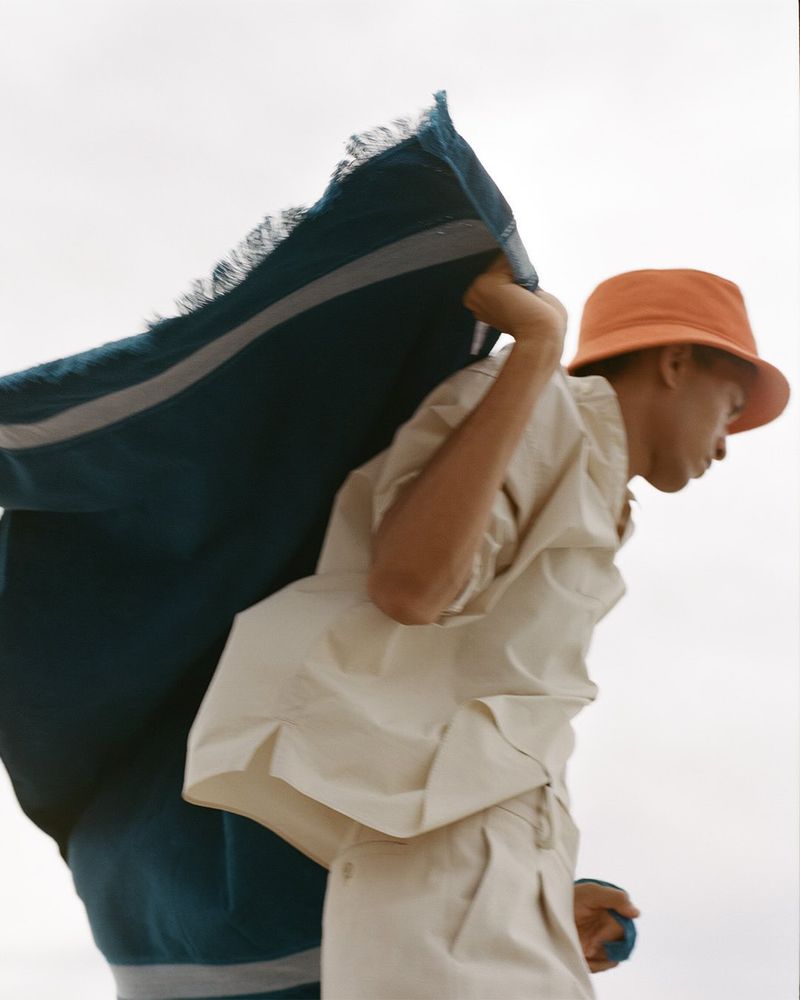 Young adult carries blue and white striped textile over his shoulder, wearing a beige outfit and an orange bucket hat in a studio shot.