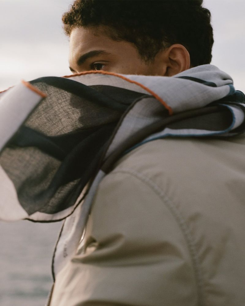 Close-up of a young adult with dark hair and tan jacket with a patterned scarf covering the mouth and nose, against a neutral background.