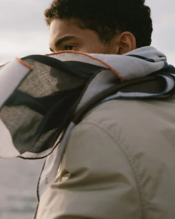 Close-up of a young adult with dark hair and tan jacket with a patterned scarf covering the mouth and nose, against a neutral background.