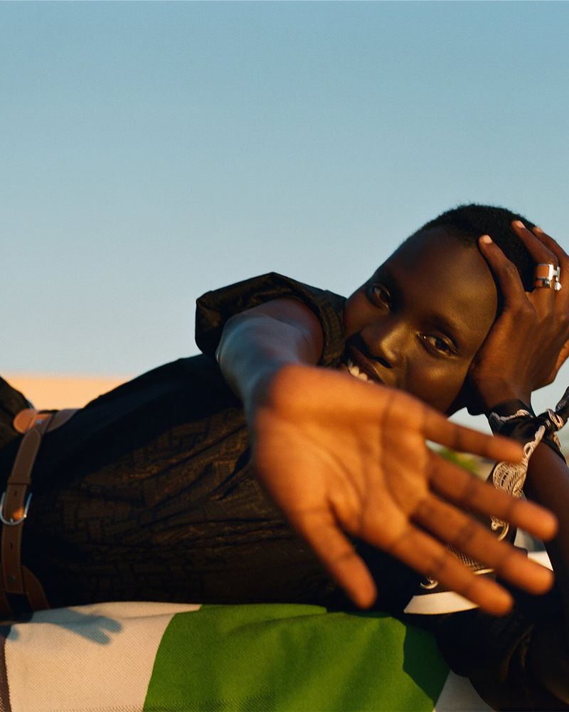Portrait of a smiling adult male laying on a striped blanket with his hand raised toward the viewer against a bright blue sky.