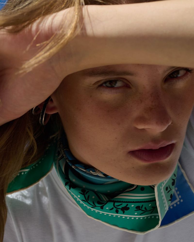 Close-up portrait of a young adult woman with a patterned headscarf shielding her eyes from the sun in a natural light setting.