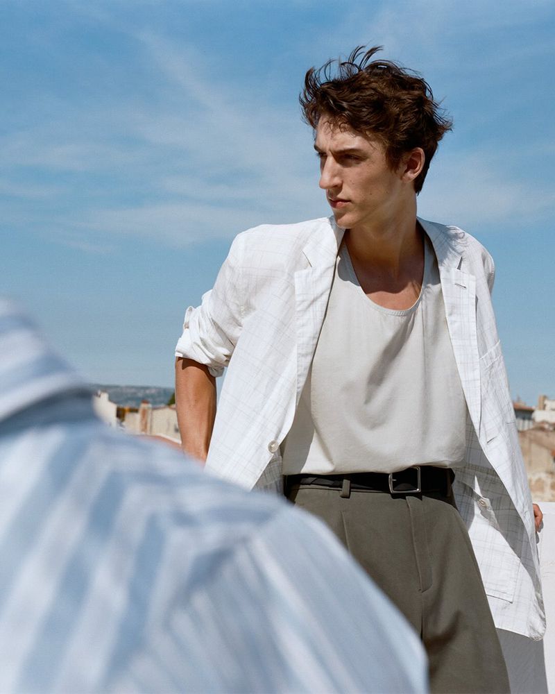 Fashion shot of a young adult male wearing a light linen jacket, t-shirt, and dark pants, standing outside on a sunny day.