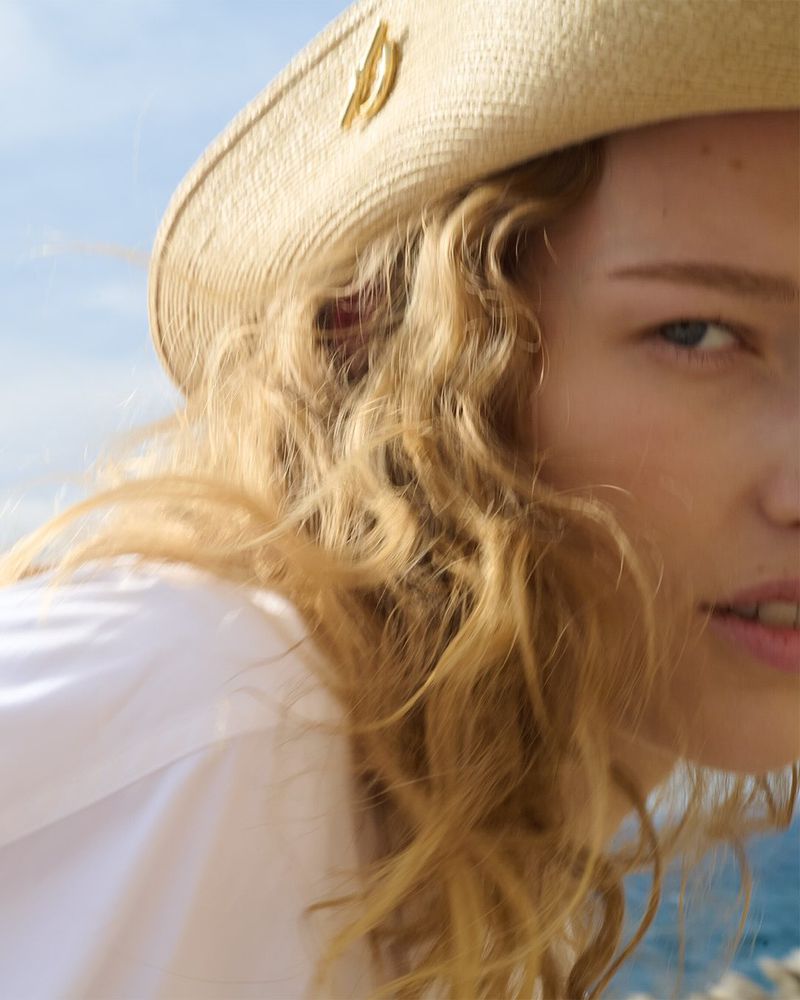 Close-up of a woman with fair skin and curly strawberry blonde hair, wearing a straw hat and a white shirt, with a blue sky background.