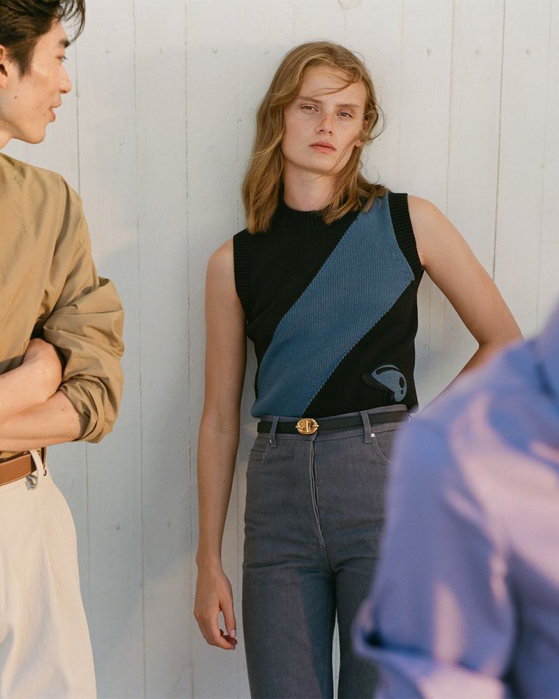Fashion editorial image: a woman in a black and blue top and grey pants poses with two other people on a white paneled background.