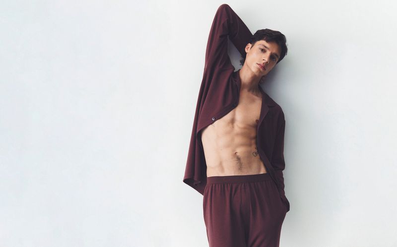 Handsome, toned adult male poses against a white wall, wearing an open maroon shirt and matching maroon pants in a fashion setting.