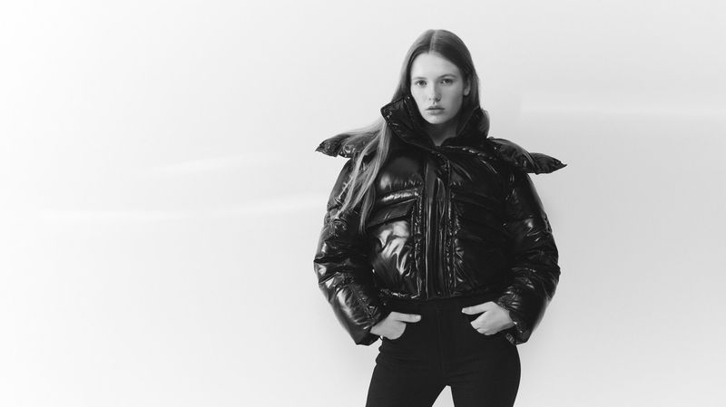 Monochrome portrait of a woman in a glossy black puffer jacket and black pants posing in front of a white backdrop, hands in pockets.
