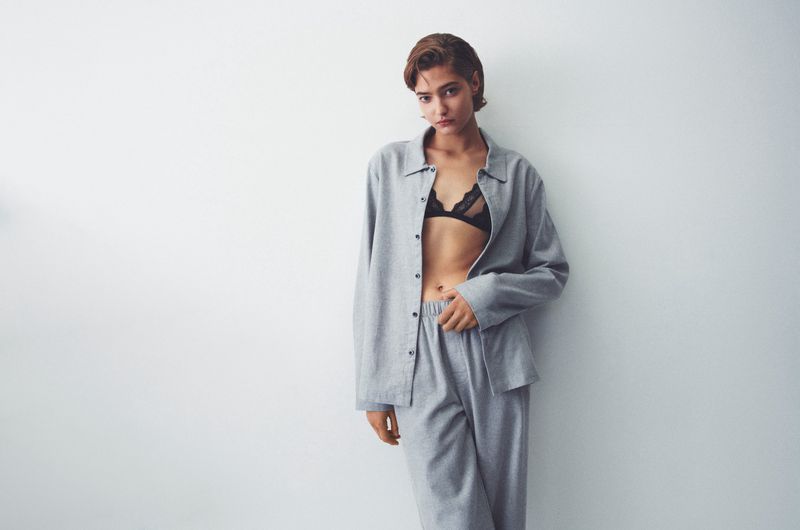 A woman with short hair in a grey lounge set and black lace bra stands against a plain white wall in a fashion studio shot.