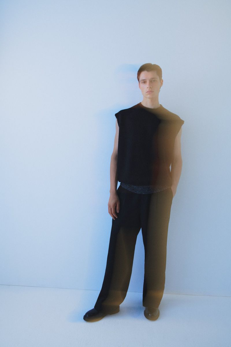 A young adult stands in a photo studio wearing a sleeveless black top and black slacks against a bright white backdrop.