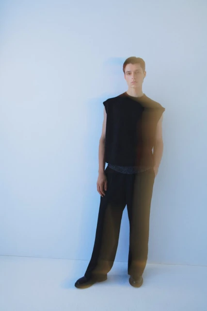 A young adult stands in a photo studio wearing a sleeveless black top and black slacks against a bright white backdrop.