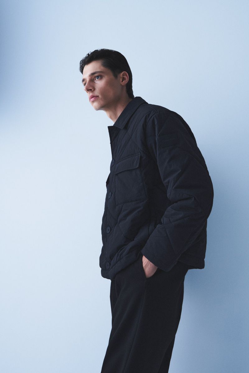 An adult male wearing a black quilted jacket and black pants is posing against a light blue background in a studio setting.