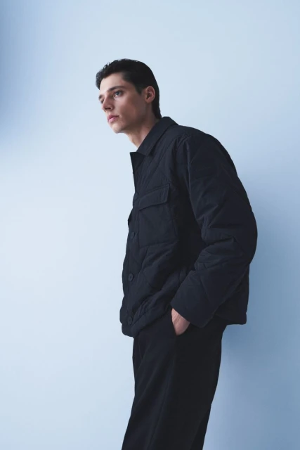 An adult male wearing a black quilted jacket and black pants is posing against a light blue background in a studio setting.