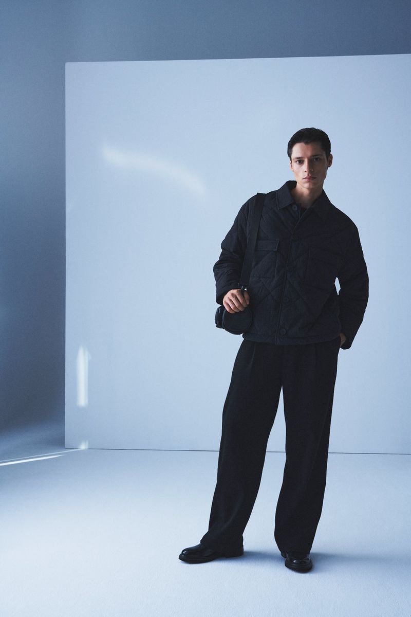 A slender man wearing a black jacket, black trousers, and a small black bag stands in a blue-toned studio with soft, diffused lighting.
