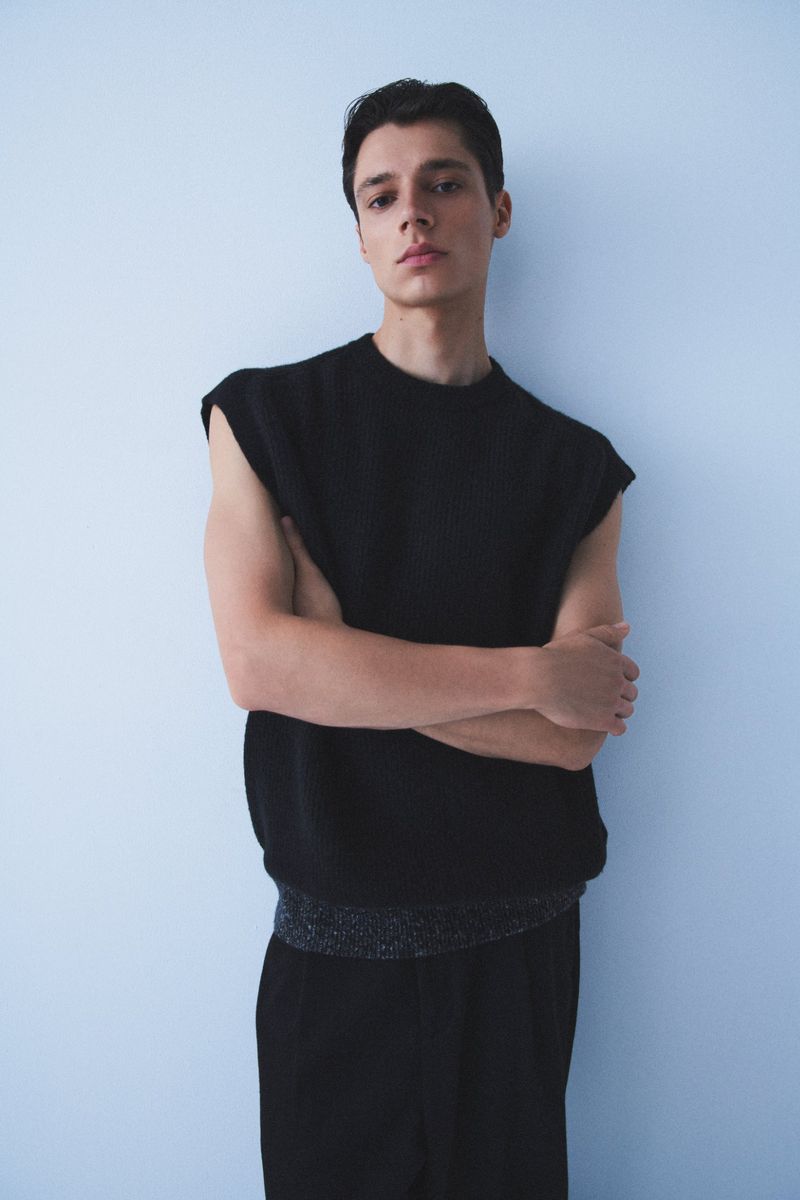 Indoor studio shot of a young adult male in a black sleeveless knit top and black pants against a light blue wall, posing with arms crossed.