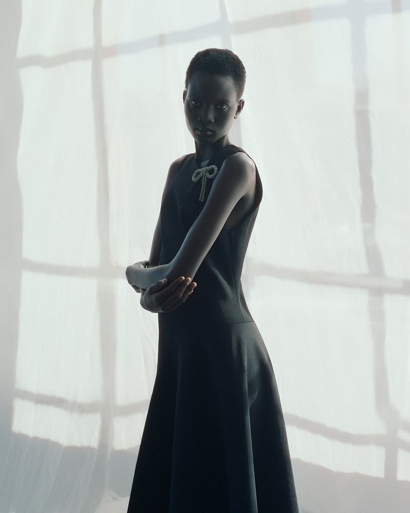 Dark-skinned woman poses in a sleeveless black dress, arms crossed, set against a softly lit white backdrop, creating a fashion portrait.