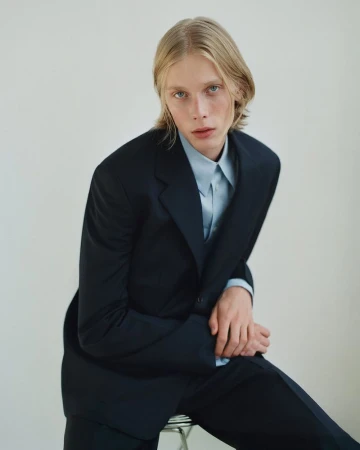 Young man with blonde hair wearing a dark blazer and pale blue shirt seated against a light background