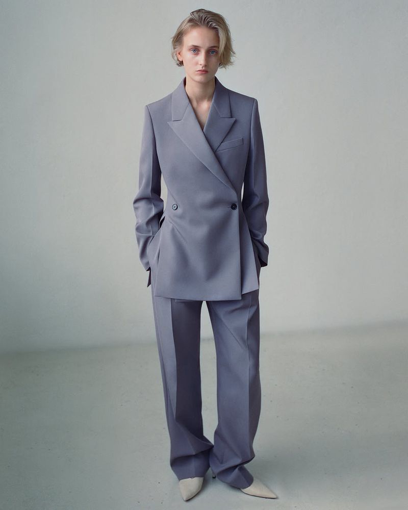 Adult woman wearing a double-breasted blazer and pants suit in a light gray shade, posing in a studio setting. Fashion photography.