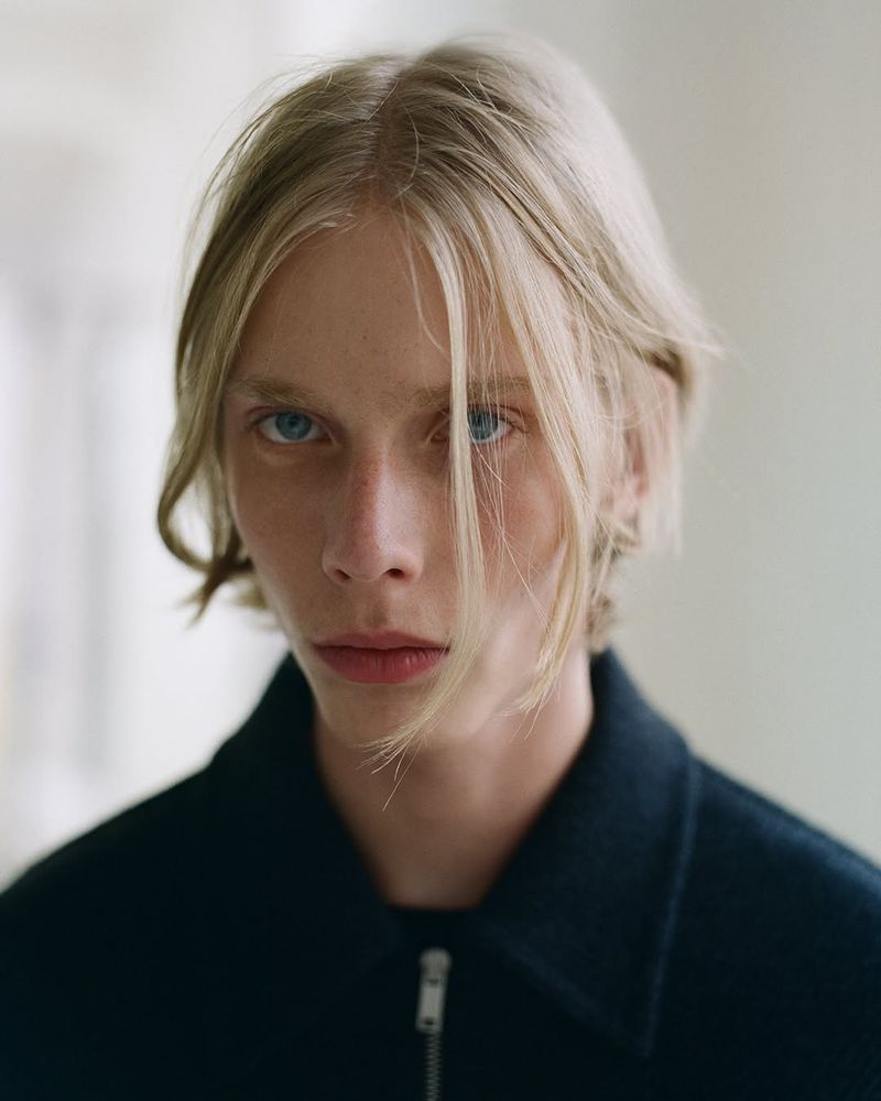Close-up portrait of a young adult male with blond hair, blue eyes, and wearing a dark blue jacket, photographed indoors.