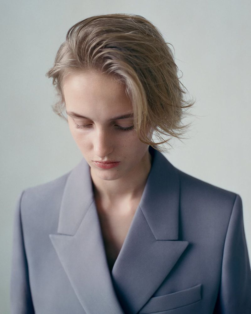 Close-up portrait of a young, fair-skinned woman with wavy blonde hair, wearing a blue-grey blazer, against a light background.