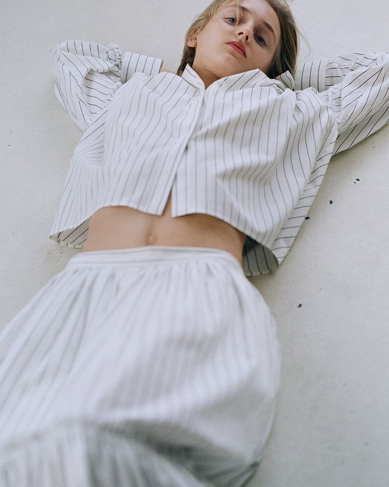 An adult female lying on a light surface wearing a striped, collared shirt and skirt with long hair and neutral facial expression.
