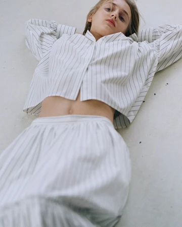 An adult female lying on a light surface wearing a striped, collared shirt and skirt with long hair and neutral facial expression.