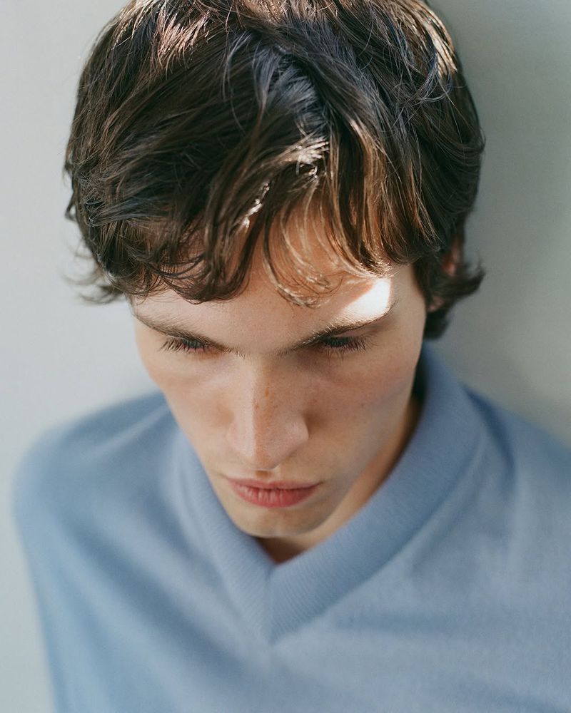 Headshot of a young adult male with dark wavy hair wearing a light blue V-neck shirt, looking down in a bright indoor setting.
