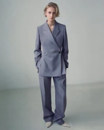 Model standing in a grey double-breasted suit jacket and matching trousers against a pale grey studio background.
