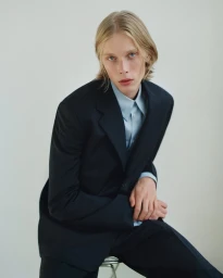 Young man with blonde hair wearing a dark blazer and pale blue shirt seated against a light background