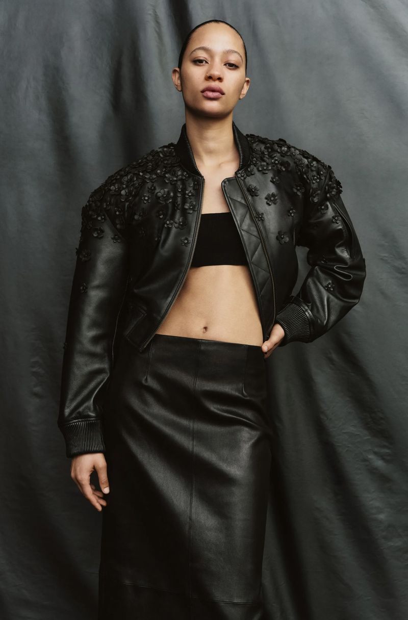 Woman in black leather jacket and skirt poses against draped fabric backdrop in studio lighting for fashion editorial shot.