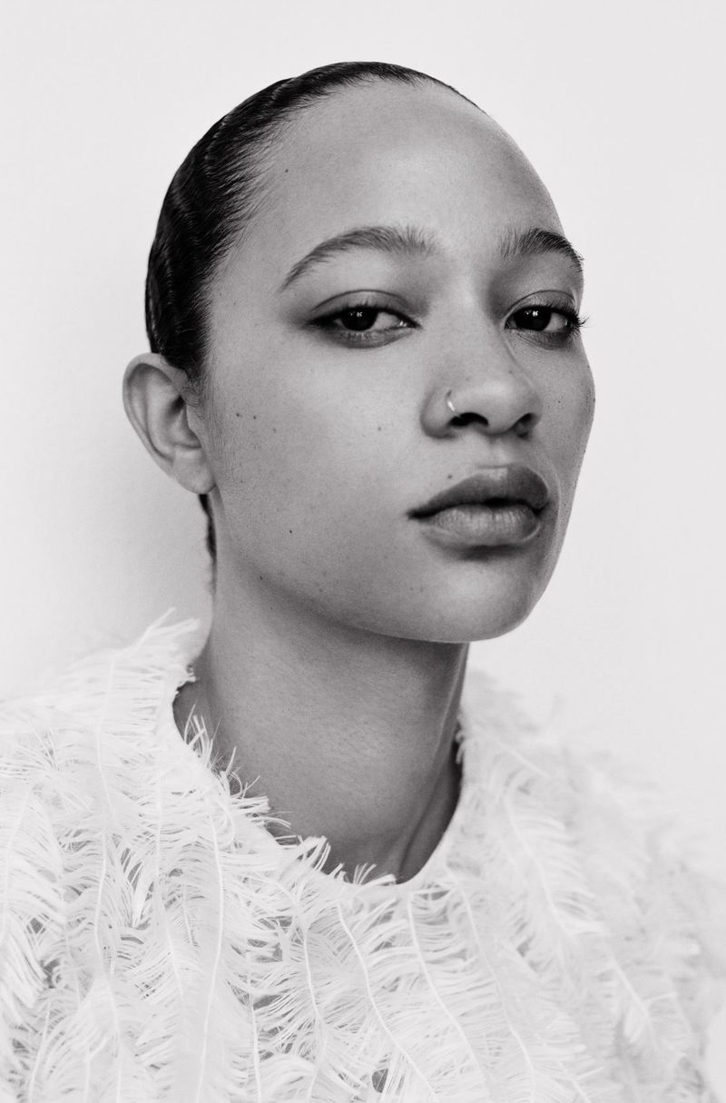 Striking black and white portrait of a young Black woman with a nose ring wearing a textured white top against a white backdrop.