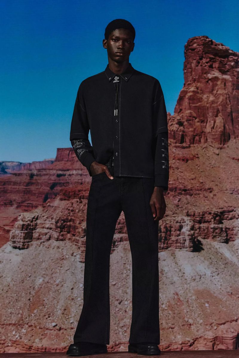 Full-length portrait of a dark-skinned male fashion model in a dark shirt and pants, set against a landscape backdrop with mesas.