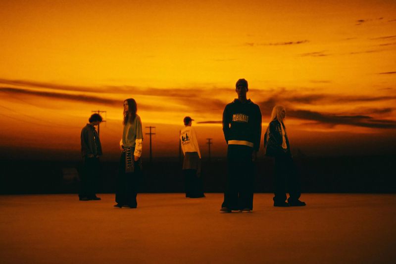 Group of five people standing in a flat landscape with a vibrant orange and yellow sunset sky, wearing casual attire.