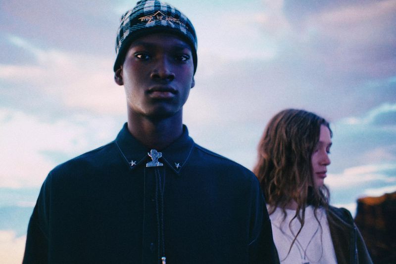 A young Black male wearing a beanie and bolo tie stands in front of a fair-skinned person and cloudy sky in an outdoor setting.