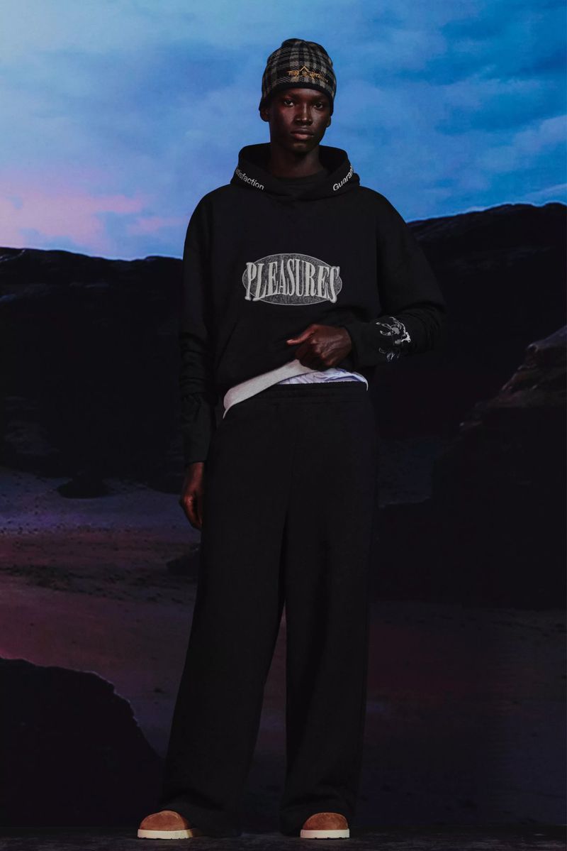 Dark-skinned adult male fashion model wears a black hoodie, wide-legged pants, beanie, and brown shoes in a studio against a dark landscape backdrop.